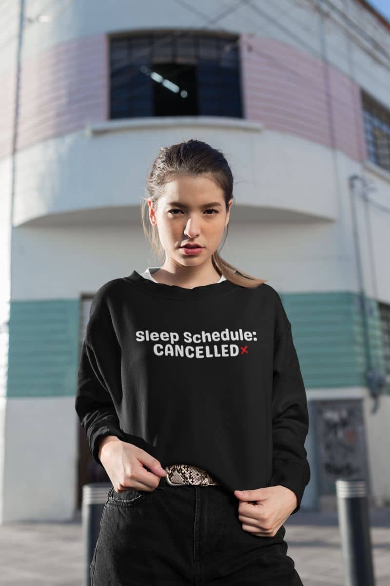 exhausted life sweater with a person wearing a black sweatshirt with 'Sleep Schedule: CANCELLED' text in front of a building.