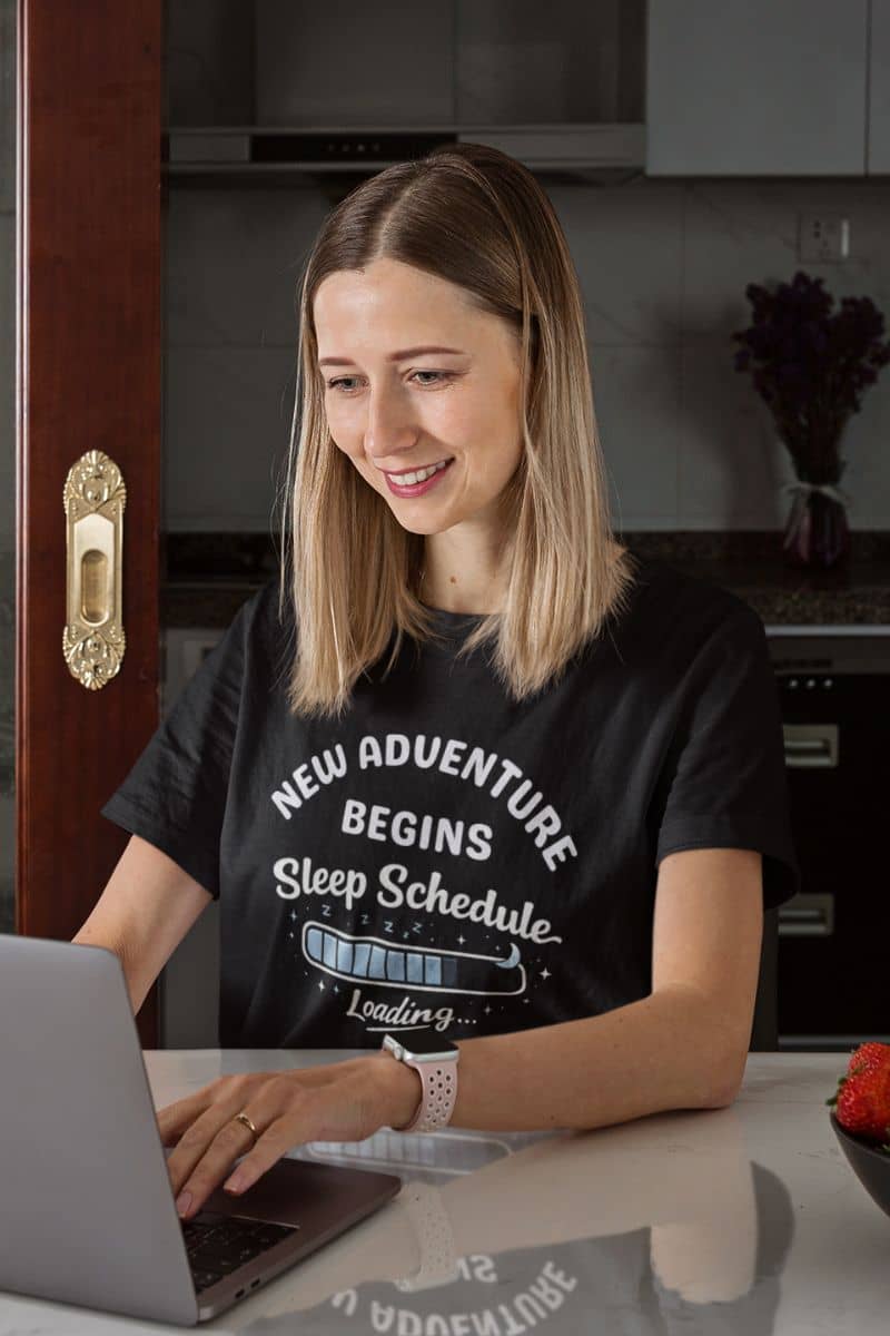 exhausted parent shirt with a woman wearing a black t-shirt with text 'New Adventure Begins, Sleep Schedule Loading…', sitting at a kitchen counter using a laptop.