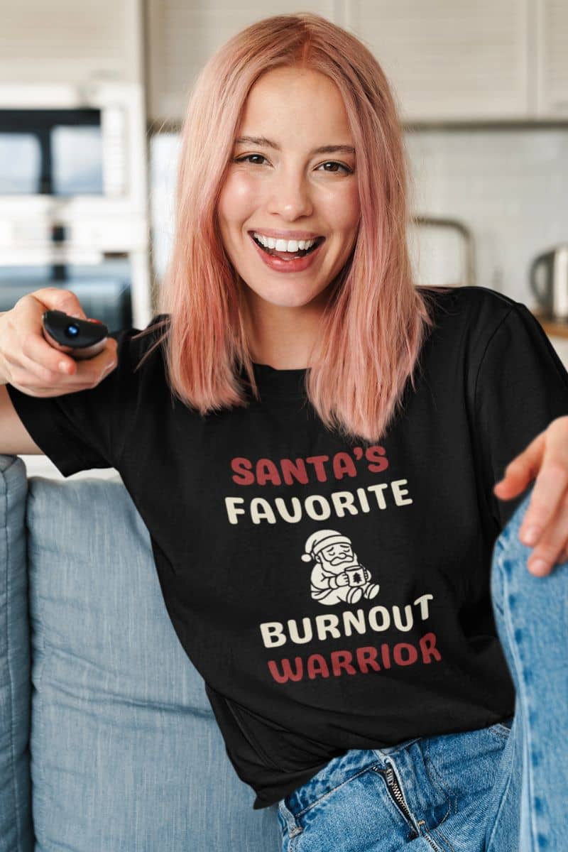 exhausted santa shirt with a person wearing a black t-shirt that reads 'Santa's Favorite Burnout Warrior' text, sitting on a couch.
