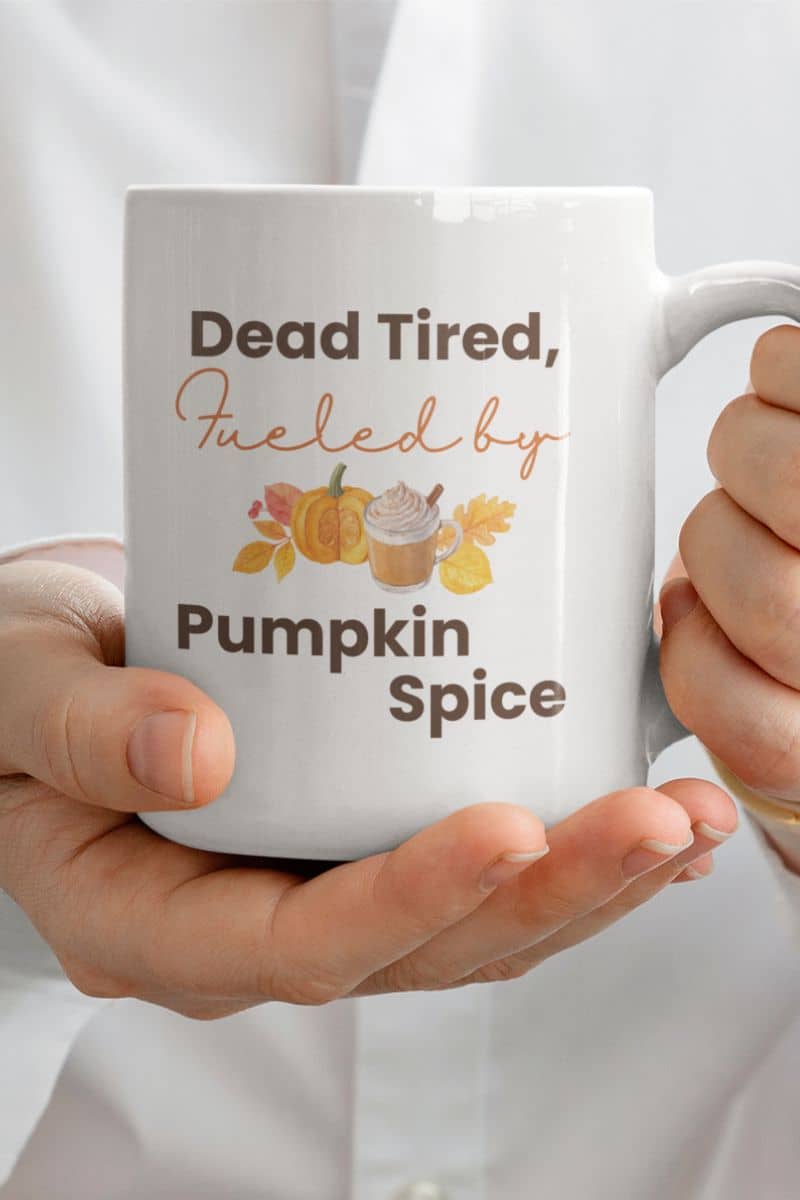 fall quote mug with a white mug with 'Dead Tired, Fueled by Pumpkin Spice' text and autumn-themed graphics held by a person.