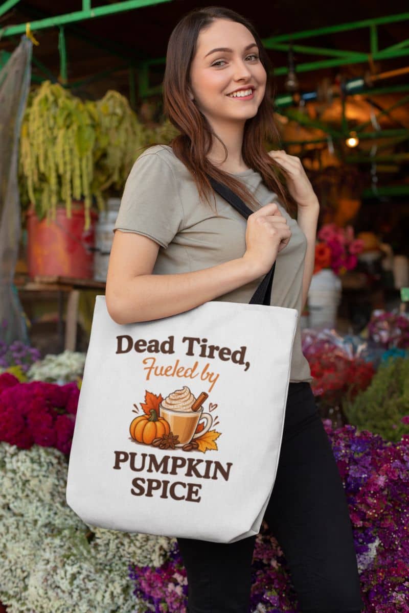 A woman carrying a white canvas tote bag with a humorous pumpkin spice latte graphic and the text 'Dead Tired, Fueled by Pumpkin Spice' in a colorful outdoor flower market setting.