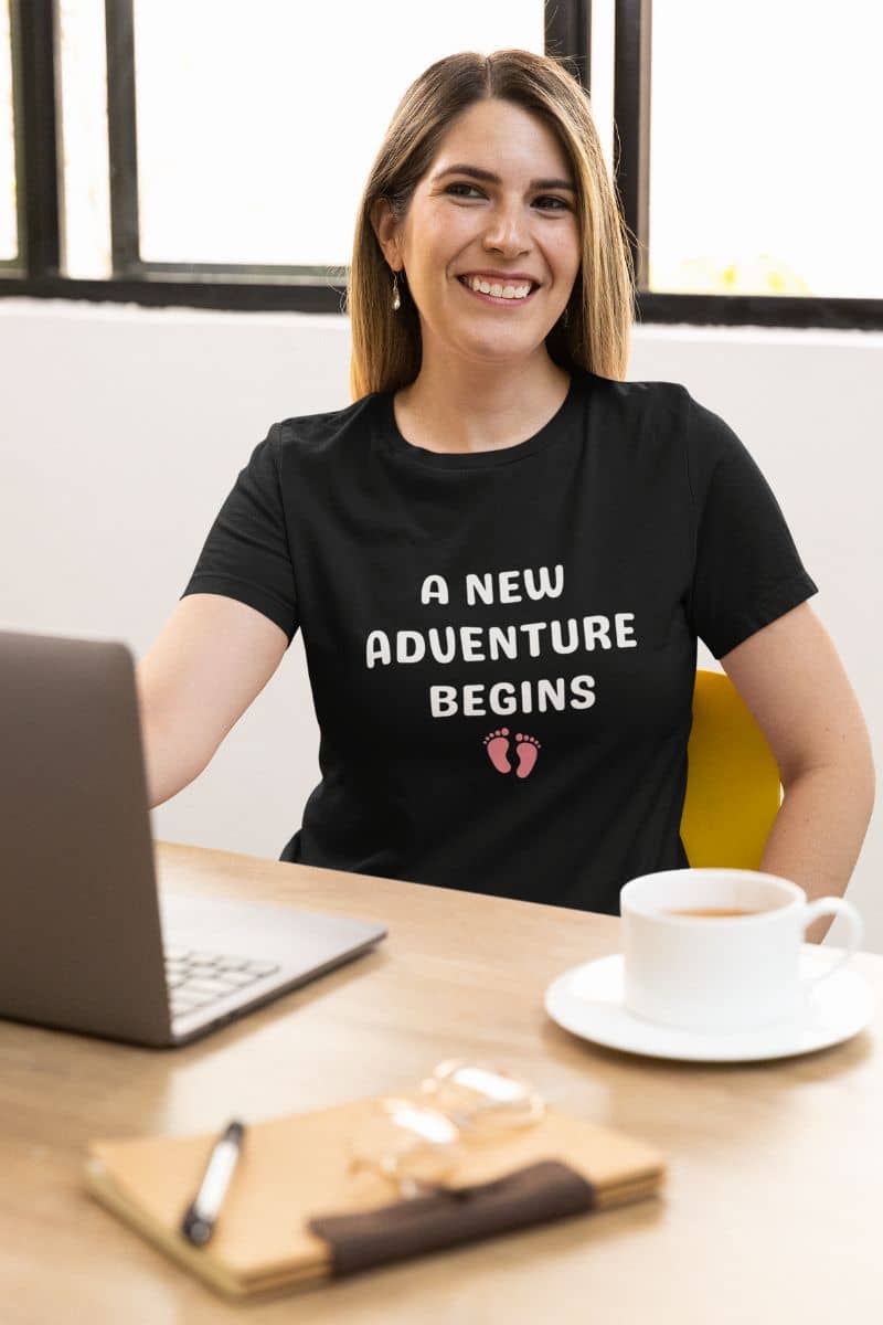 family life shirt with a woman wearing a black t-shirt with 'A New Adventure Begins' text, sitting at a desk with a laptop and coffee.