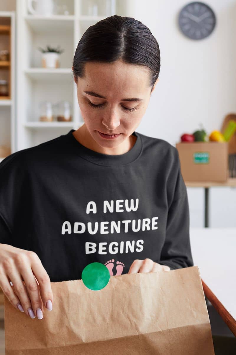family life sweatshirt with a person wearing a black sweatshirt with 'A New Adventure Begins' text, opening a brown paper bag.
