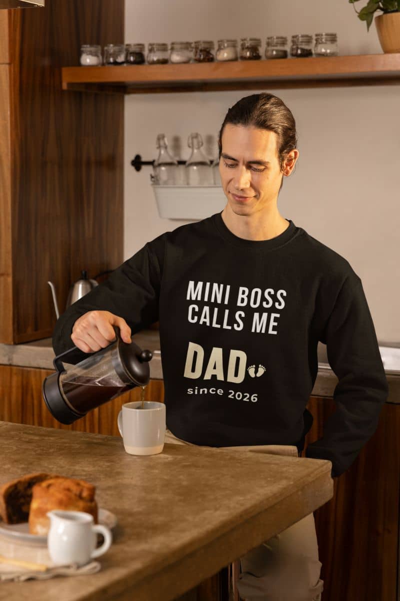 fatherhood sweatshirt with a man pouring coffee wearing a black sweatshirt with text 'Mini Boss Calls Me Dad – since 2026' in a kitchen.