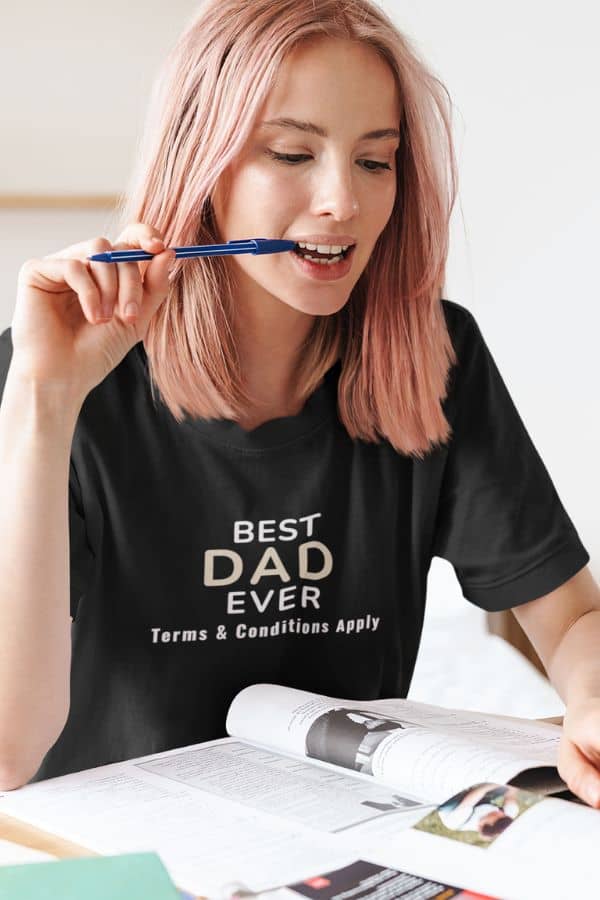 fathers day funny shirt with a person wearing a black t-shirt with 'Best Dad Ever, Terms & Conditions Apply' text, holding a pen and looking at a magazine.