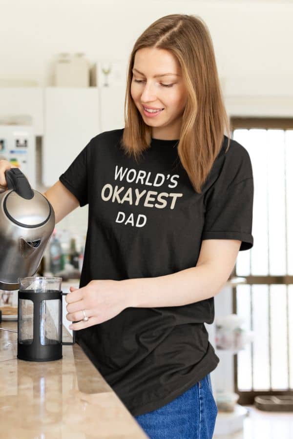 fathers day shirt with a woman in a kitchen wearing a black t-shirt with 'World's Okayest Dad' text.