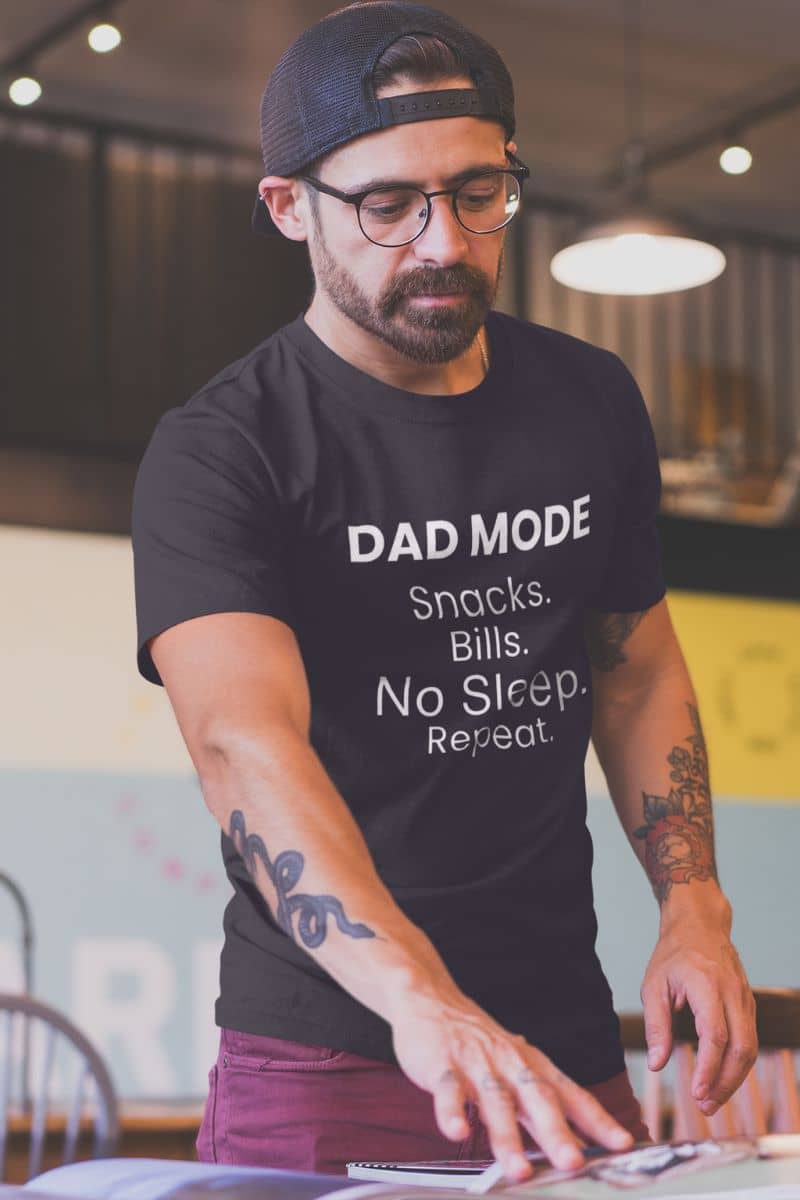 fathers day shirt with a man wearing a black t-shirt with text 'DAD MODE:  Snacks. Bills. No Sleep. Repeat.', sitting at a table.