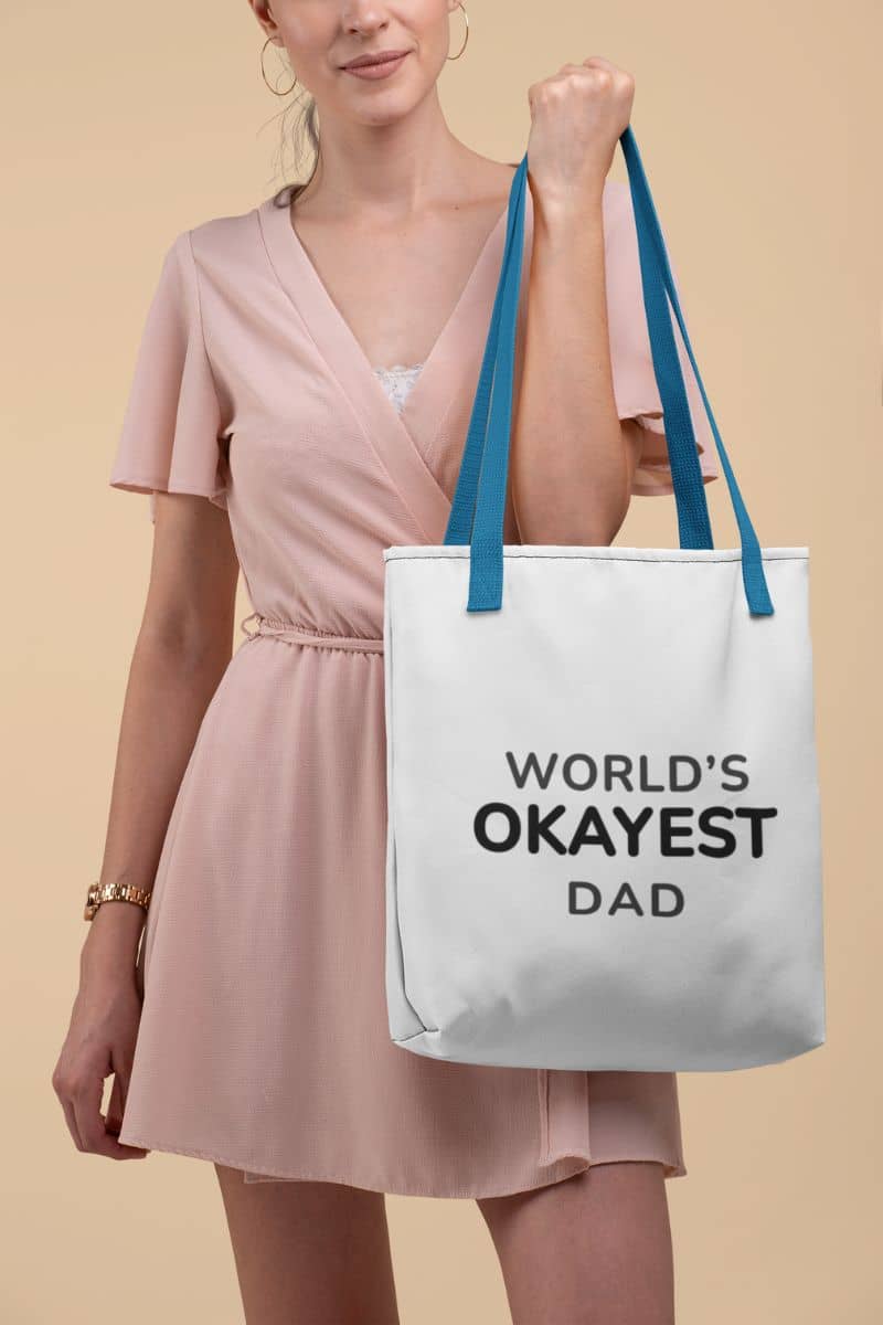father's day tote bag with a woman holding a tote bag with 'World's Okayest Dad' text against a beige background