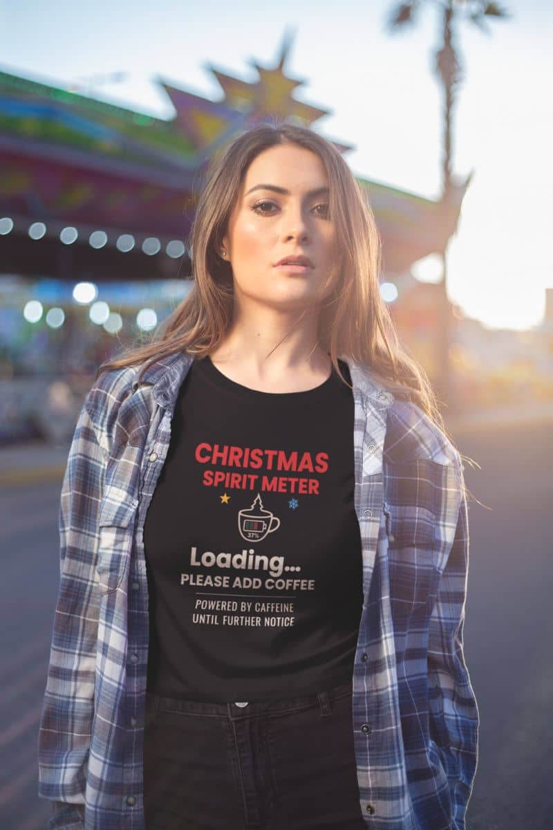 festive caffeine tee with a woman wearing a black t-shirt with 'Christmas Spirit Meter, Loading…37%. Please add coffee. Powered by caffeine until further notice' text in a festive setting.