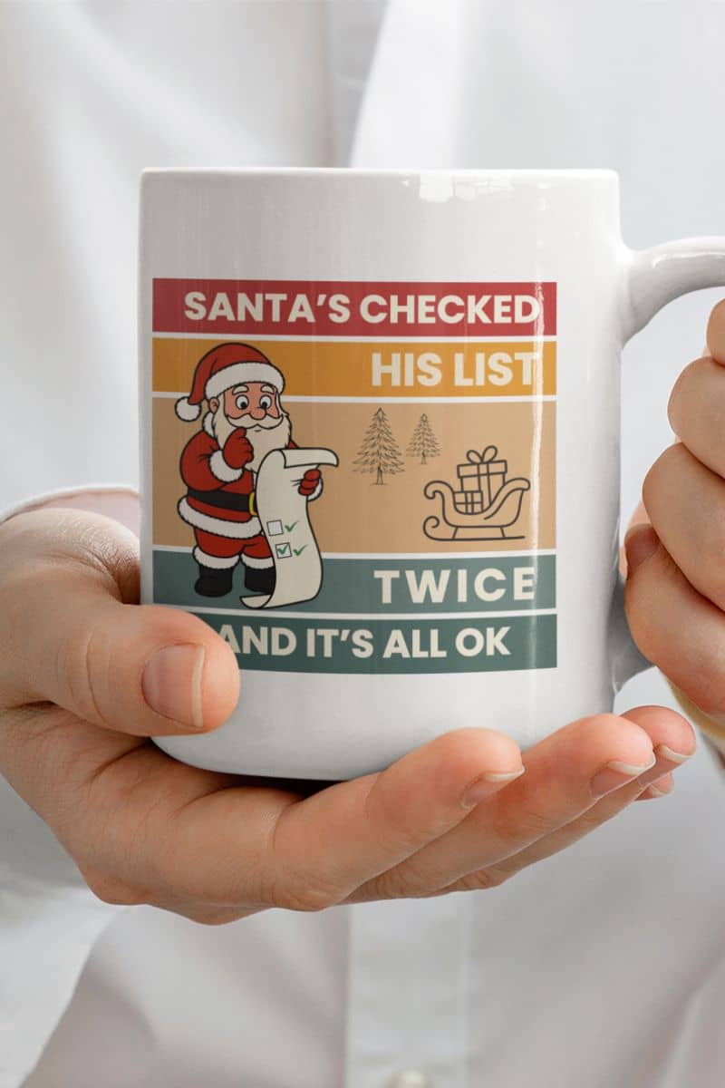 festive ceramic mug with a mug with Santa Claus design and text 'Santa's Checked His List Twice And It's All Ok' held by a person