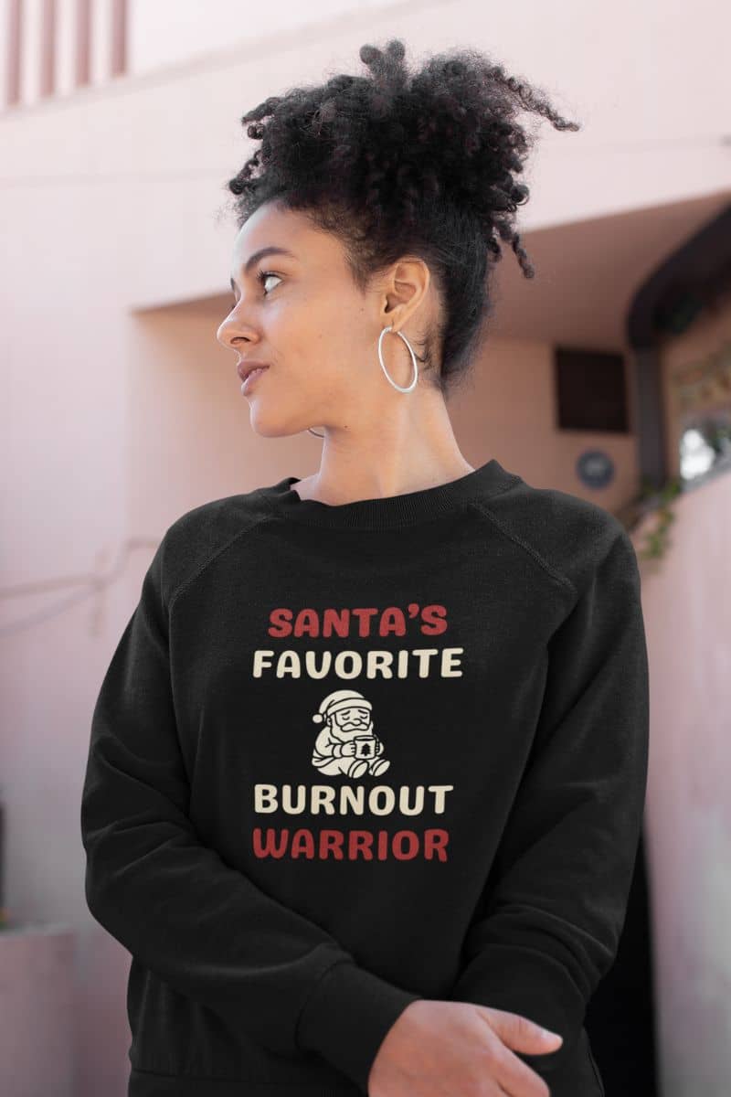 festive cozy pullover with a person wearing a black sweatshirt with text 'Santa's Favorite Burnout Warrior' in an indoor setting.