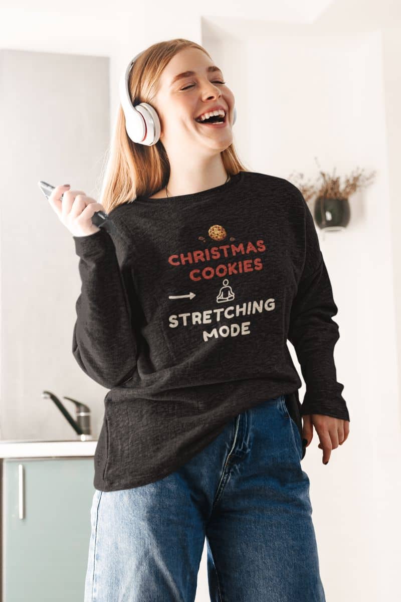 festive mental health sweatshirt with a person wearing a sweater with text 'Christmas Cookies, Stretching Mode', holding a phone, and smiling in a kitchen.