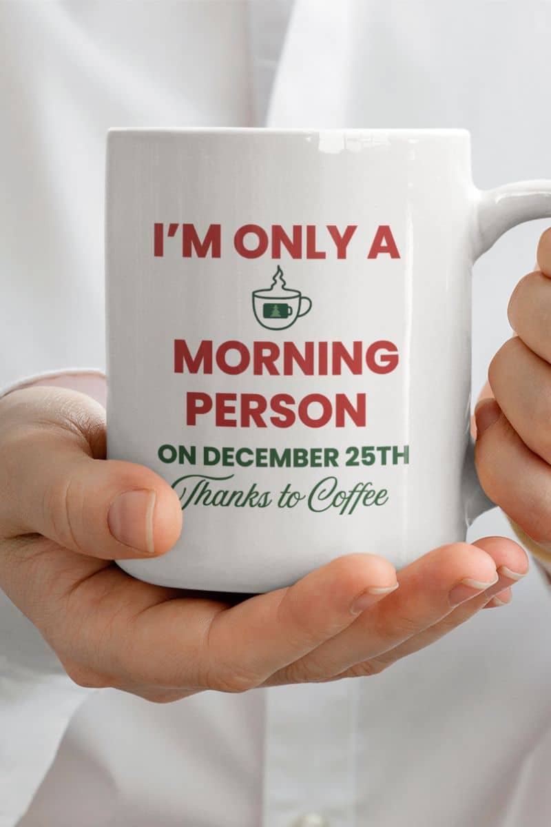 festive morning coffee mug with a white mug with humorous text 'I’m Only a Morning Person on December 25th' held by a person, on a white background