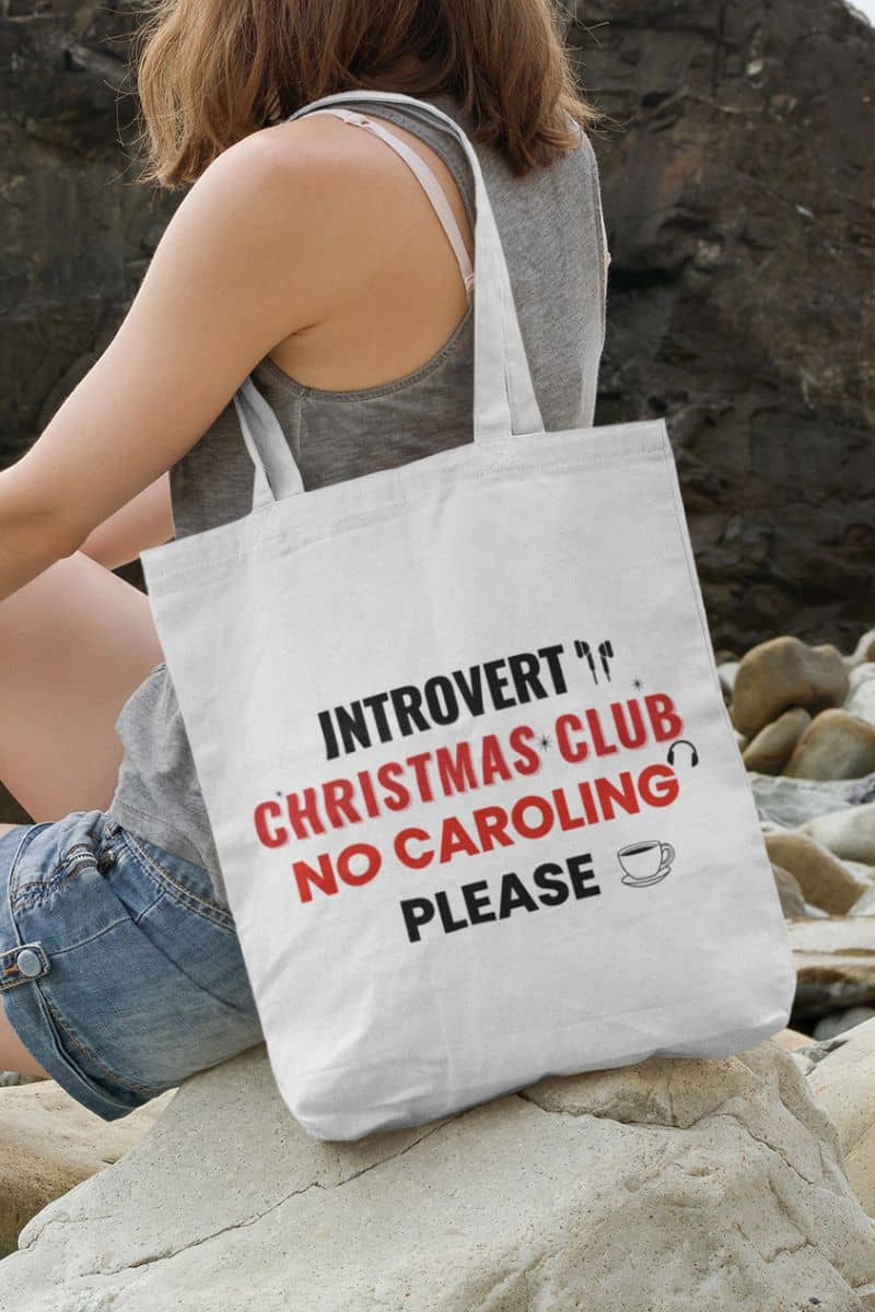 festive shopping tote with a person sitting on rocks with a tote bag featuring text 'Introvert Christmas Club, No Caroling Please'.