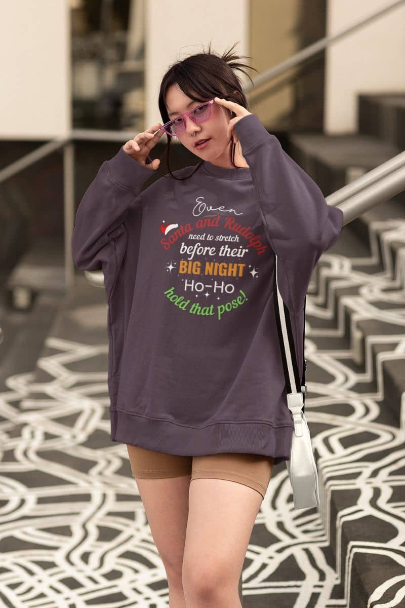 Festive winter sweater featuring a woman in a purple oversized sweatshirt with the text “Even Santa & Rudolph Need to Stretch Before Their Big Night. Ho-Ho-Hold That Pose!”, posing with glasses