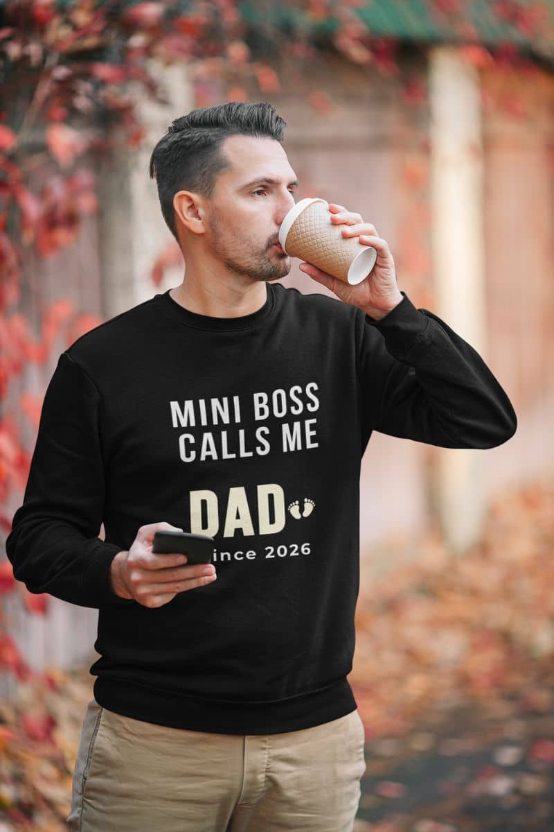 first time dad sweatshirt with a man wearing a black sweatshirt with text 'Mini Boss Calls Me Dad – since 2026', drinking from a paper cup outdoors.