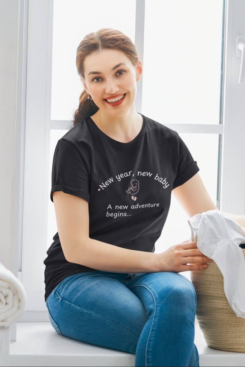 First time parents shirt worn by a woman sitting in a laundry room, holding a basket of clothes and wearing a black t-shirt with the text “New Year, New Baby. A new adventure begins…”