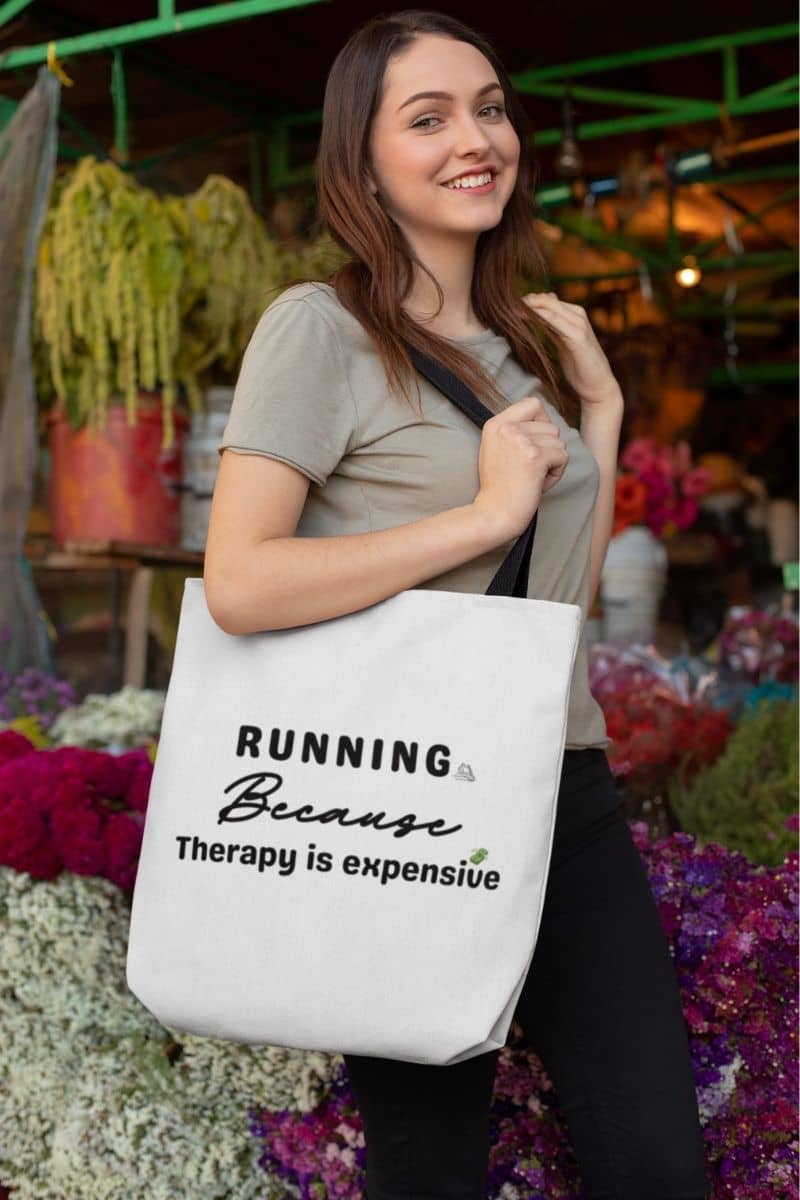 Fitness quote tote bag worn by a woman standing in front of a flower shop, featuring the text “Running because therapy is expensive.”