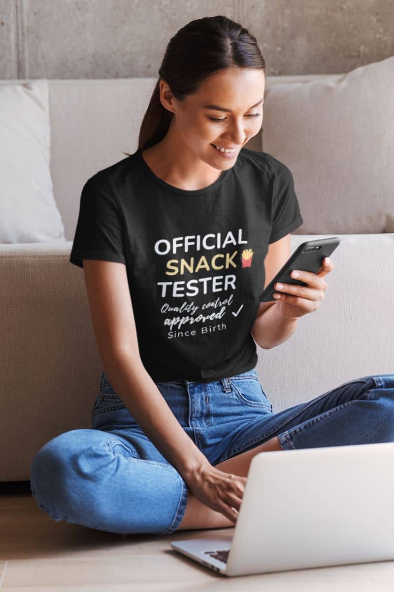 food humor shirt with a woman wearing a black t-shirt with 'Official Snack Tester – Quality Control Approved – Since Birth' text, using a phone on a couch.