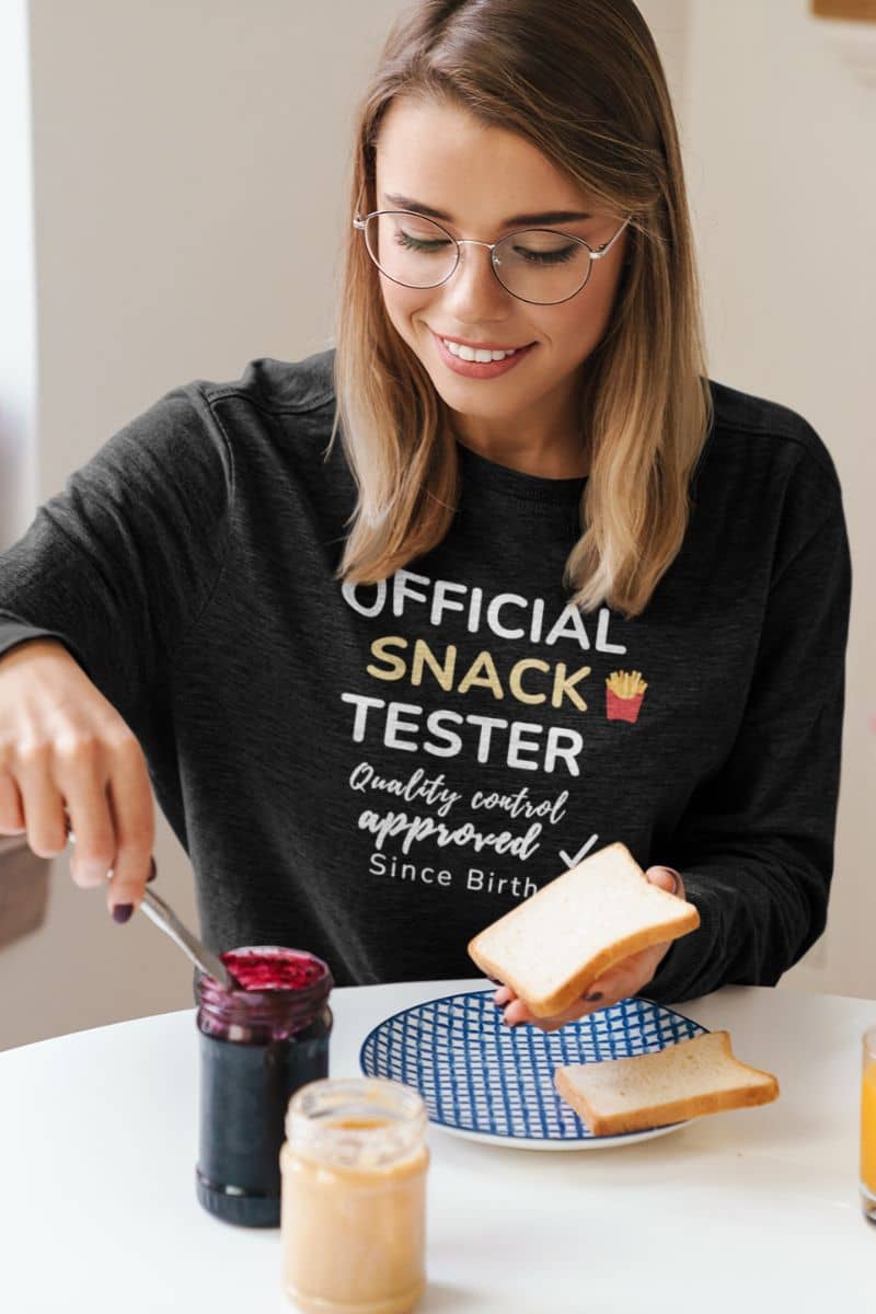 food sweatshirts with a woman wearing a t-shirt with text 'Official Snack Tester – Quality Control Approved – Since Birth', preparing a snack at a table.