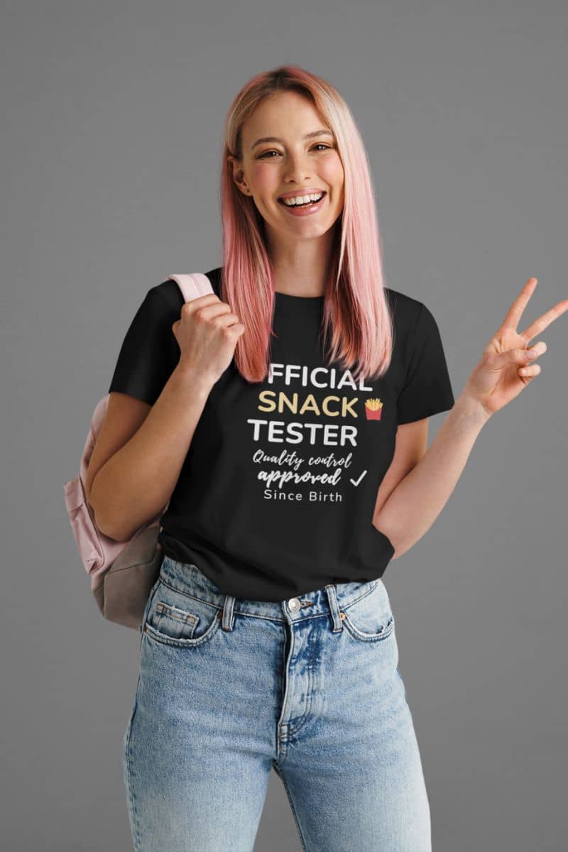 foodie t shirts with a person wearing a black t-shirt featuring the text 'Official Snack Tester – Quality Control Approved – Since Birth', holding a pink backpack on a gray background