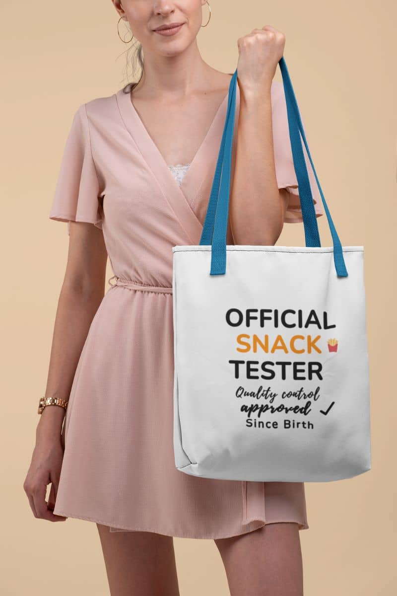 foodie tote bag with a woman holding a tote bag with text 'Official Snack Tester – Quality Control Approved – Since Birth' against a beige background