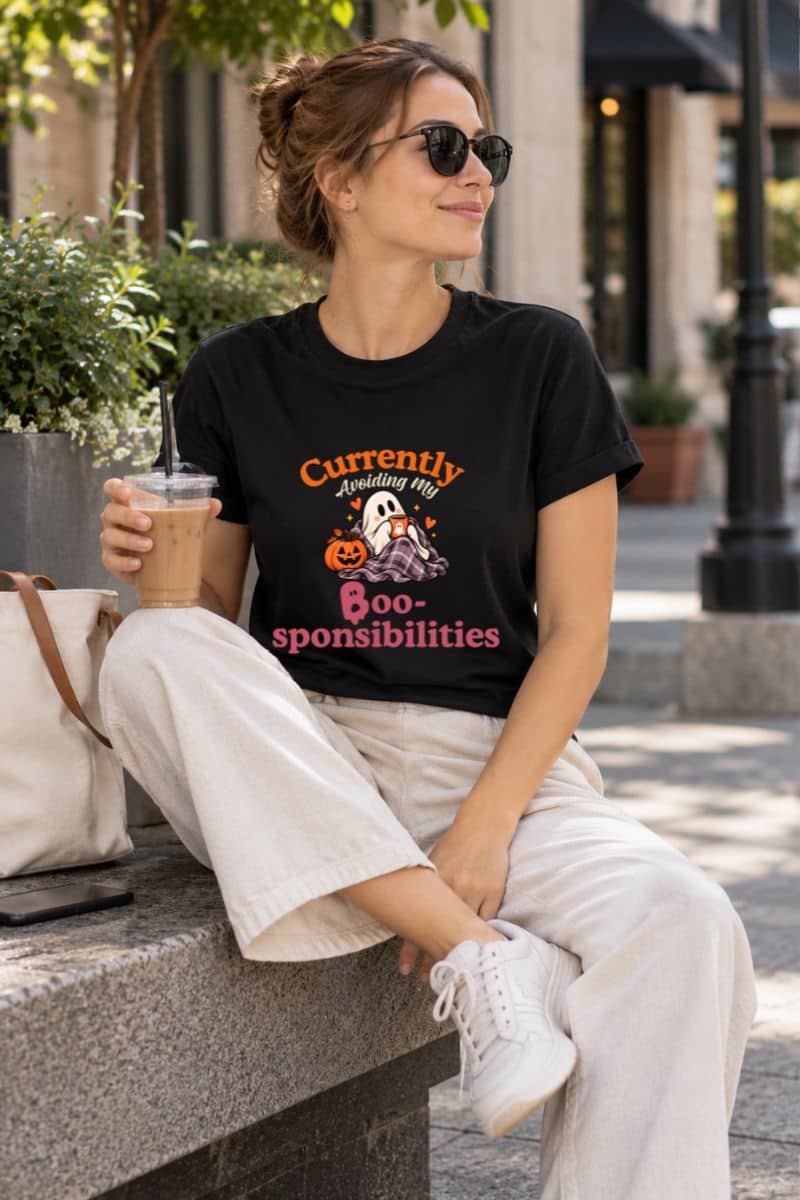 A woman sitting outdoors on a stone bench wearing a funny Halloween shirt that reads 'Currently Avoiding My Boo-sponsibilities' with a ghost and pumpkin illustration. She is also wearing white pants, white sneakers, and holding an iced coffee.