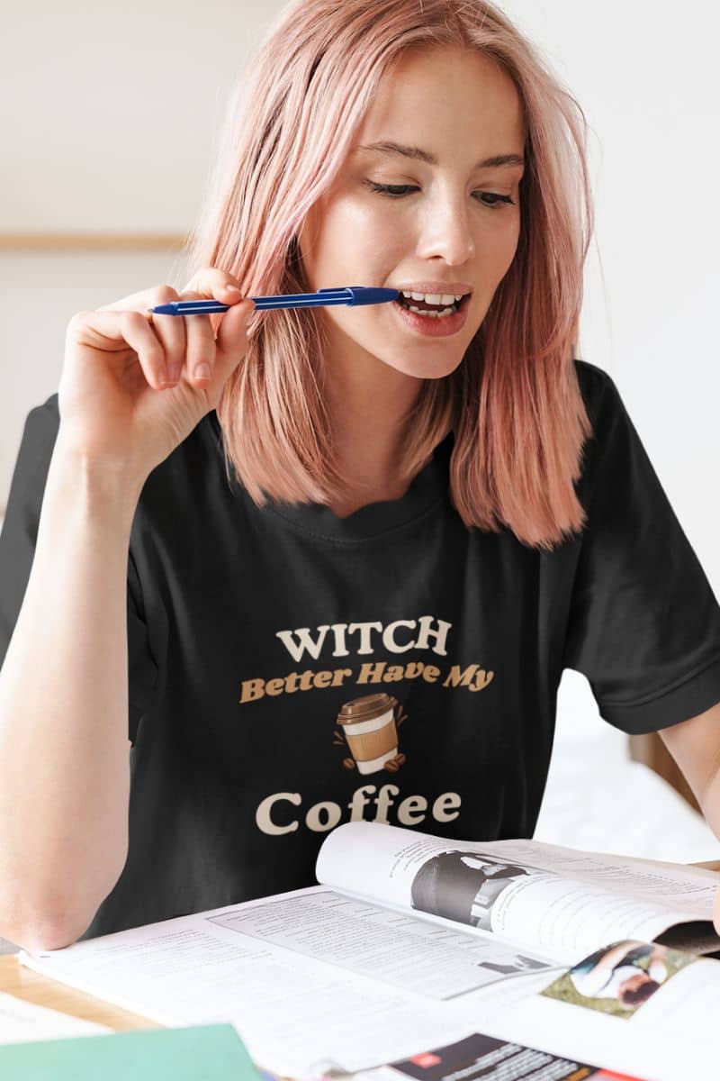 funny Halloween shirt with a woman wearing a black t-shirt featuring the text 'Witch Better Have My Coffee'. She is holding a blue pen to her mouth, with papers open in front of her.