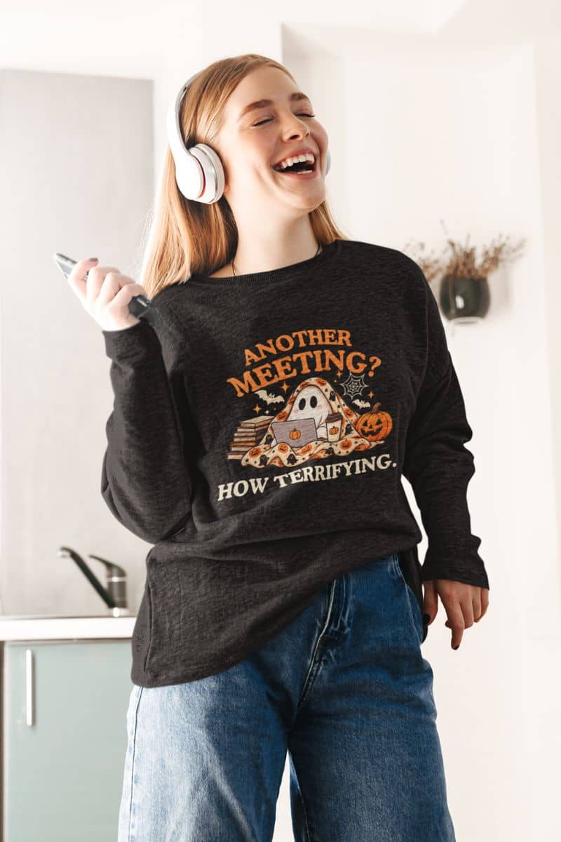 A woman wearing a funny Halloween sweatshirt featuring the text 'Another Meeting? How Terrifying.' with a ghost standing in the kitchen