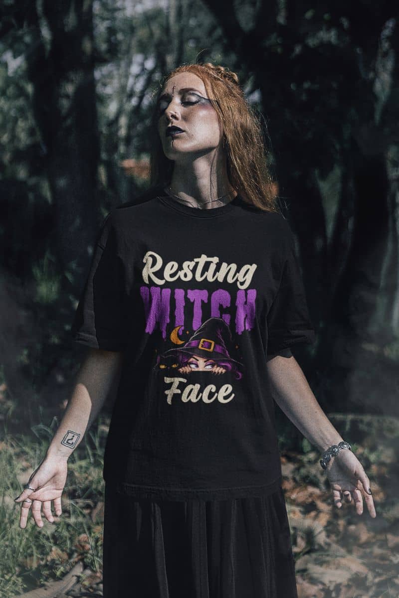 A funny Halloween tee with that reads 'Resting Witch Face' in cream and purple lettering with a witch hat llustration, worn by a woman with red hair standing in a dark forest 