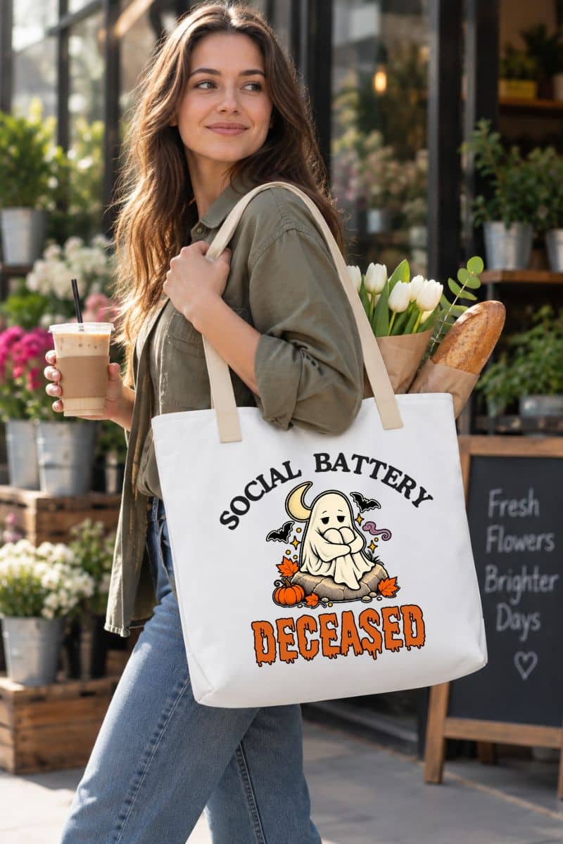 A woman carrying a funny Halloween tote with a Halloween graphic that reads 'Social Battery: Deceased' in black and orange text, featuring a ghost, moon, bats, and pumpkins.