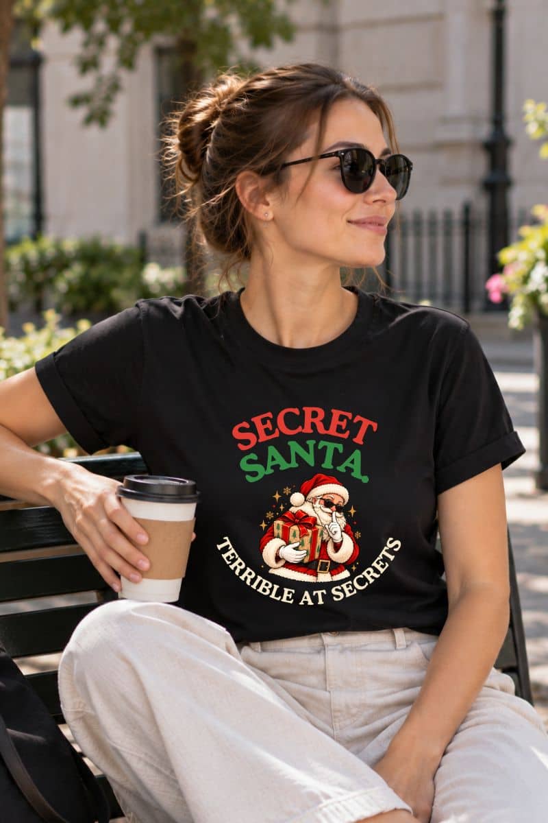 Funny Xmas shirts featuring the “Secret Santa. Terrible at Secrets.” design on a black T-shirt worn by a woman sitting outdoors with coffee.