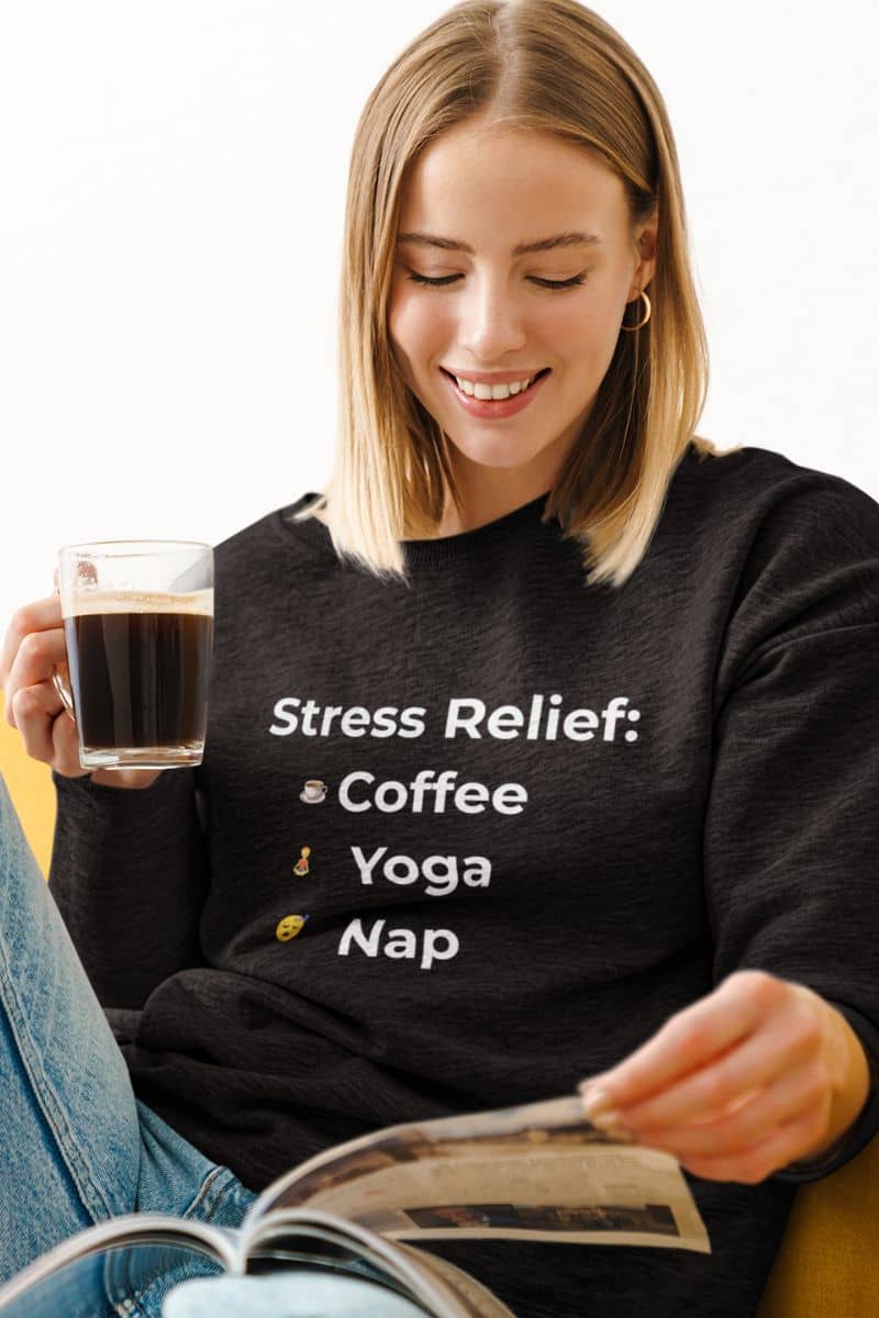 funny anxiety relief pullover with a woman wearing a black sweatshirt with 'Stress Relief: Coffee Yoga Nap' text, holding a glass of coffee and reading a newspaper.