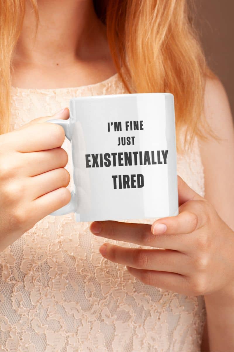 funny burnout coffee mug with a person holding a mug with text 'I'm fine just existentially tired' against a neutral background