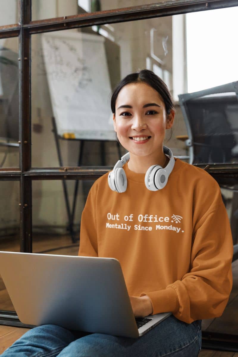 funny burnout sweater with a person wearing a mustard yellow sweatshirt with text 'Out of Office. Mentally Since Monday', sitting with a laptop in an office setting.