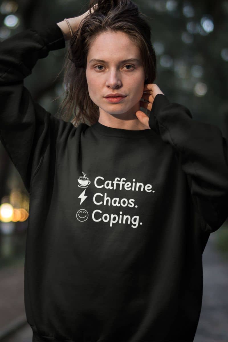funny burnout sweatshirt with a person wearing a black sweatshirt with text 'Caffeine. Chaos. Coping.' against a blurred outdoor background.