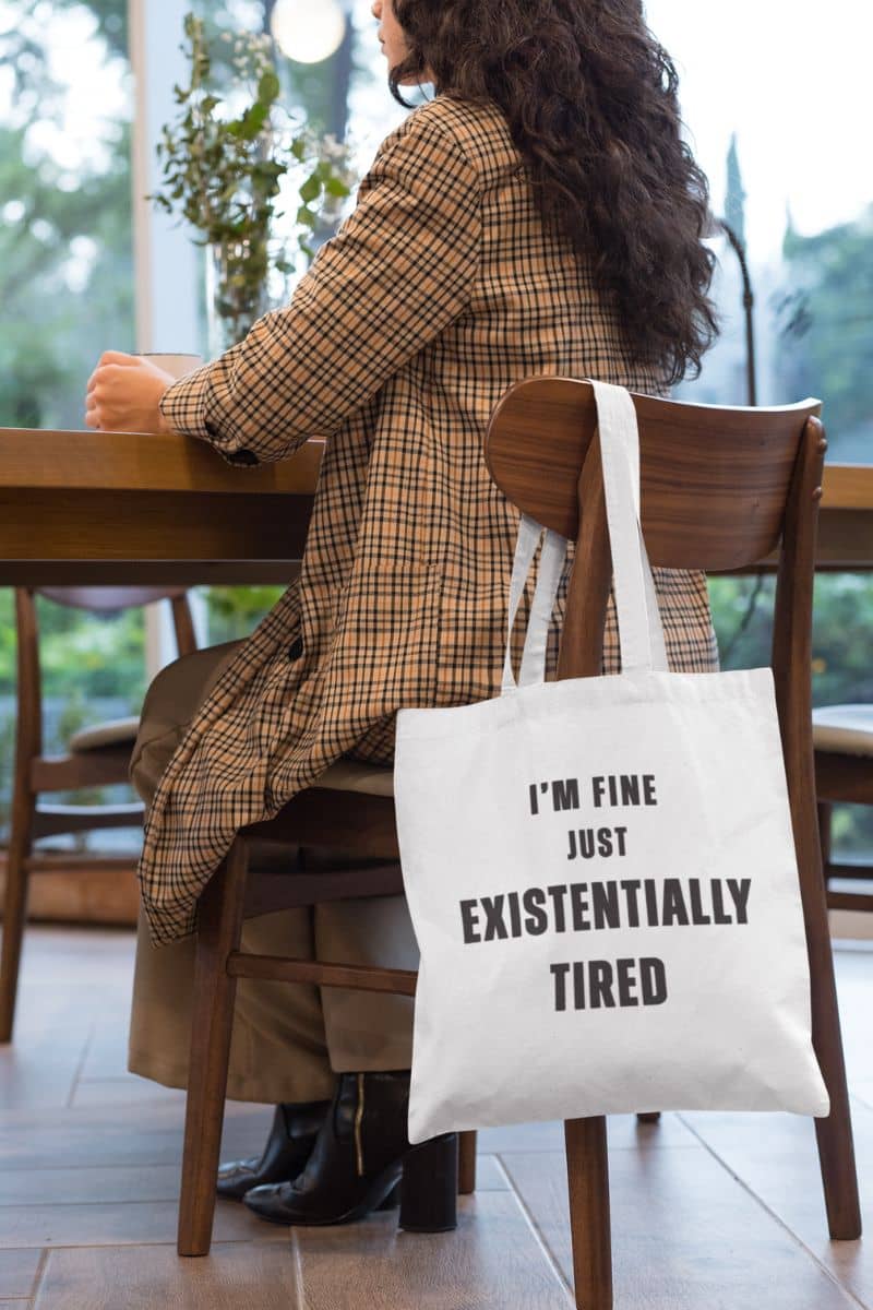 funny burnout tote bag with a person sitting at a table with a tote bag that reads 'I'm fine just existentially tired'.