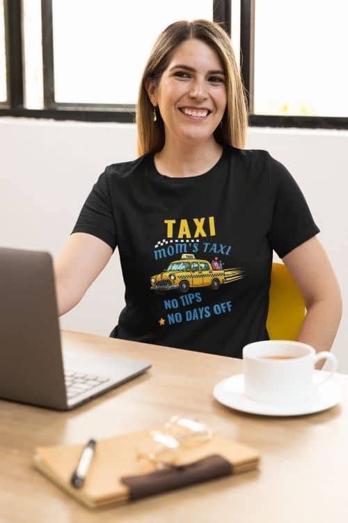 funny busy mom shirt with a woman wearing a black t-shirt with a taxi graphic and text 'Taxi, Mom’s Taxi – No tips, no days off', sitting at a desk with a laptop and coffee.