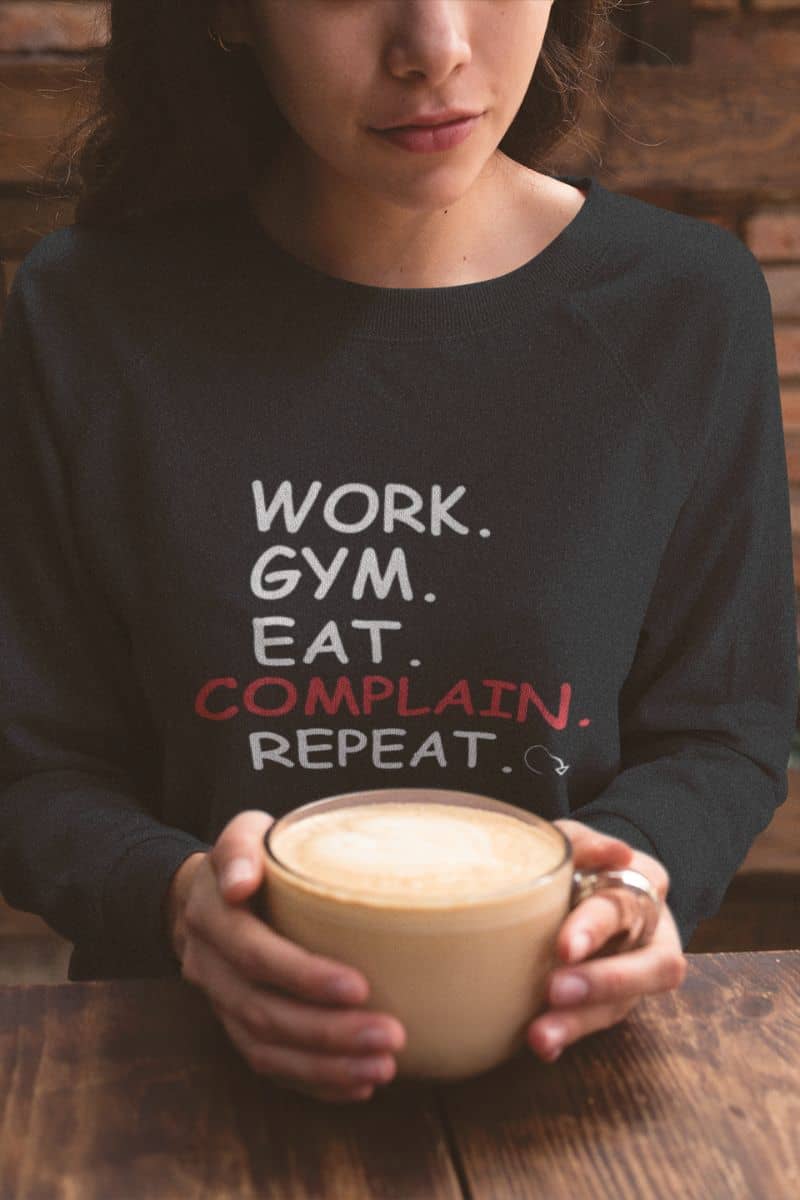 funny casual sweater with a person wearing a black sweatshirt with text 'Work. Gym. Eat. Complain. Repeat', holding a cup of coffee.