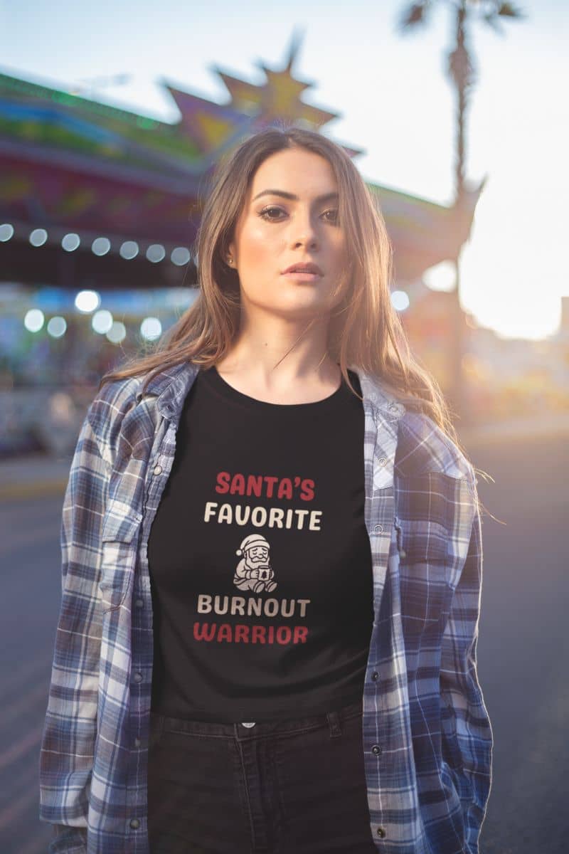 funny christmas burnout shirt with a woman wearing a black t-shirt with text 'Santa’s Favorite Burnt Out Worrior' in an outdoor setting