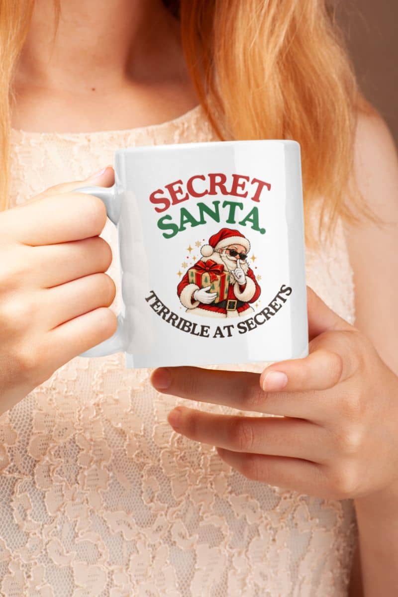 Funny Christmas mugs featuring a Secret Santa Terrible at Secrets design on a white ceramic mug held by a woman in a cream lace top.