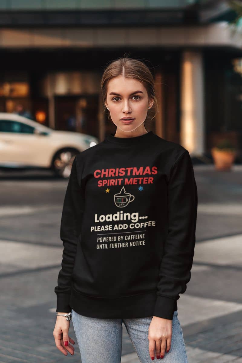 funny christmas sweater with a woman wearing a black sweatshirt with text 'Christmas Spirit Meter, Loading…37%. Please add coffee. Powered by caffeine until further notice' on a city street.