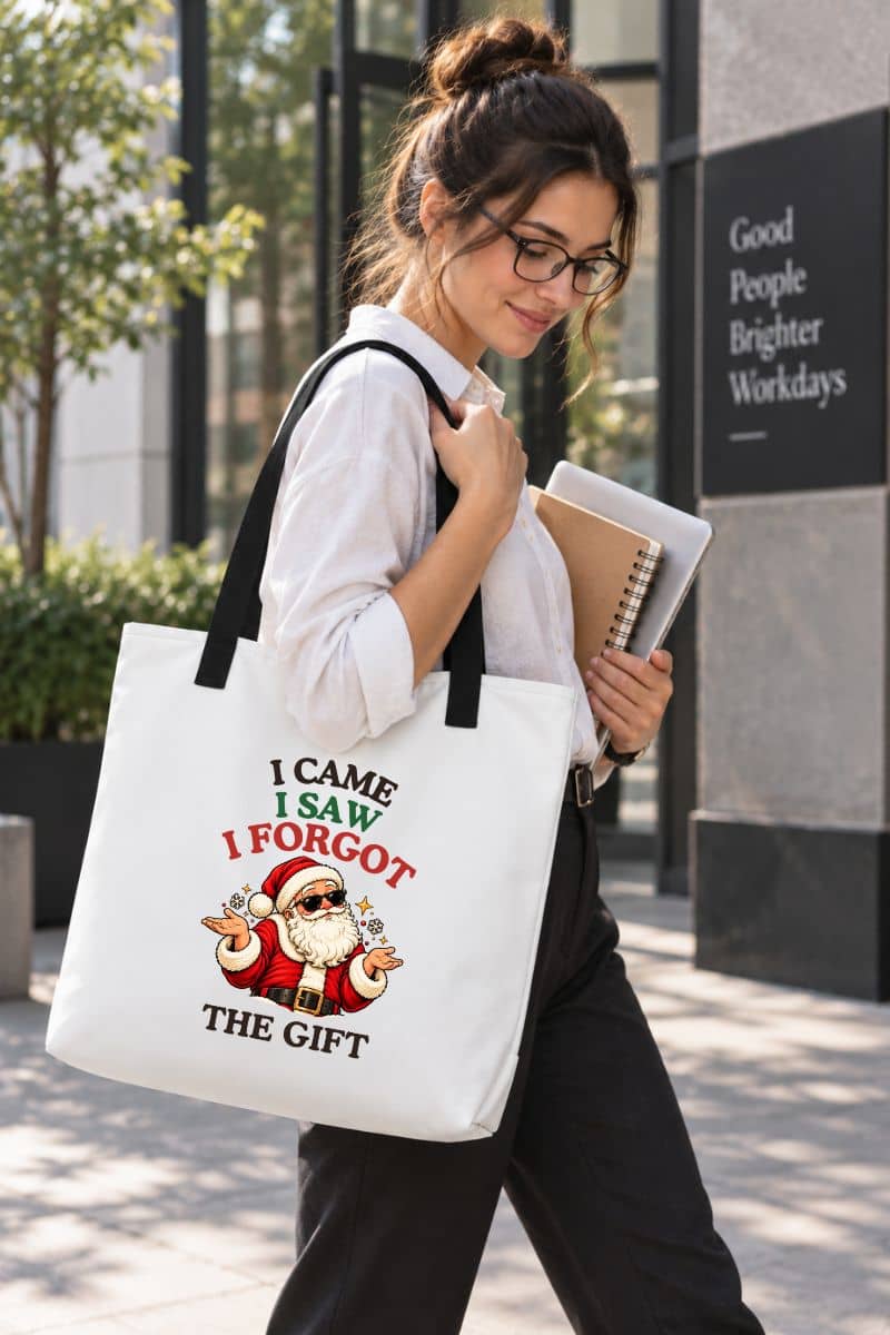 Woman wearing glasses and a white shirt carrying a funny Christmas tote bag with black handles, featuring a shrugging Santa and “I Came. I Saw. I Forgot the Gift” design.