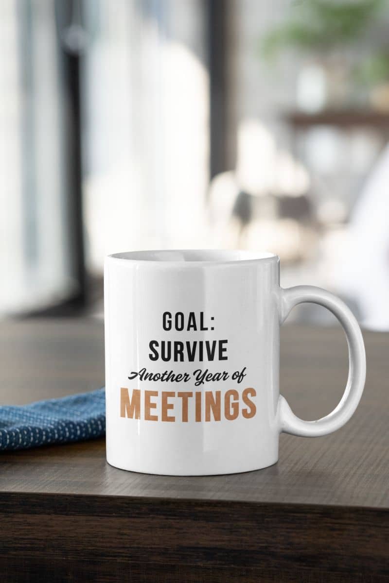 funny coffee mug for work with a mug with text 'Goal: Survive Another Year of Meetings' on a wooden surface.