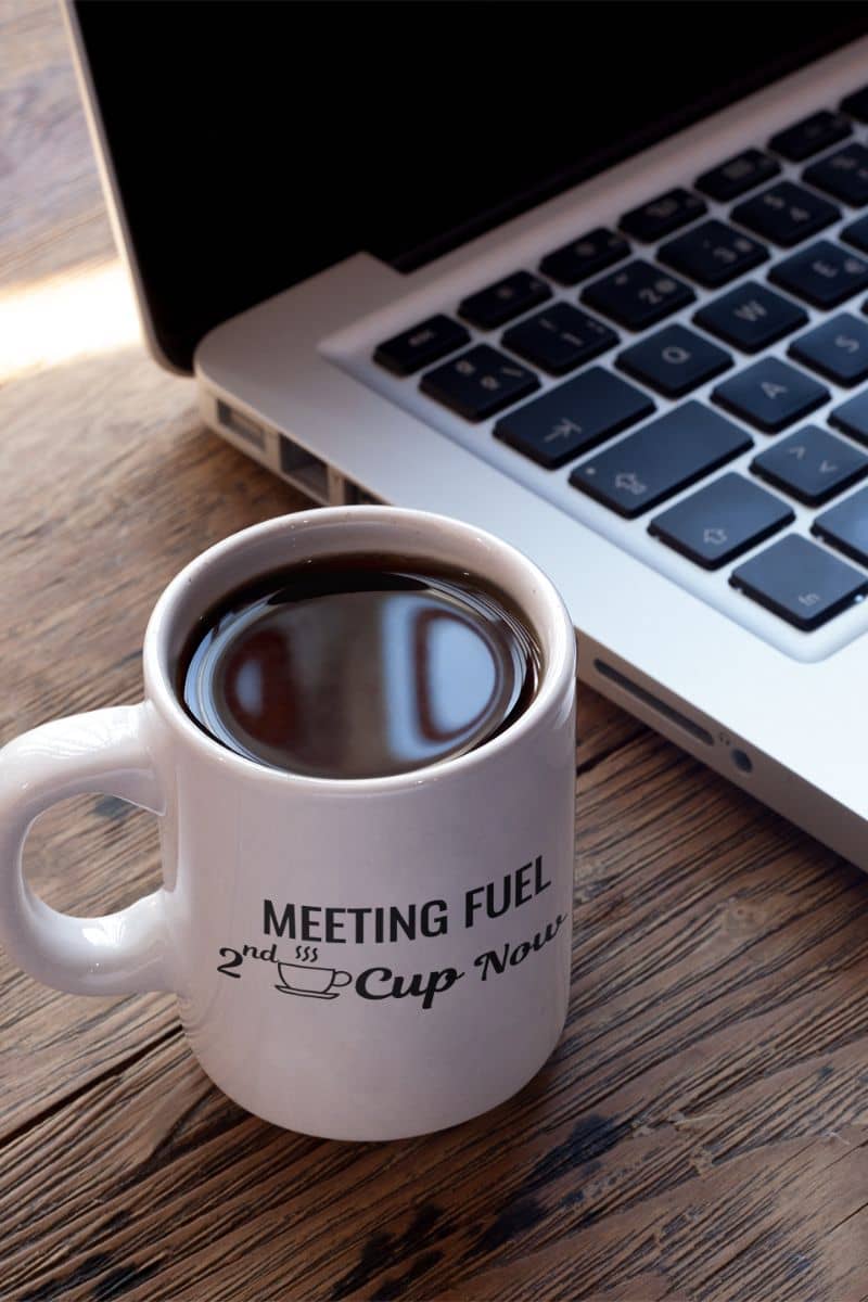funny coffee mug with a white mug with coffee and text 'Meeting Fuel: 2nd Cup Now' next to a laptop on a wooden surface