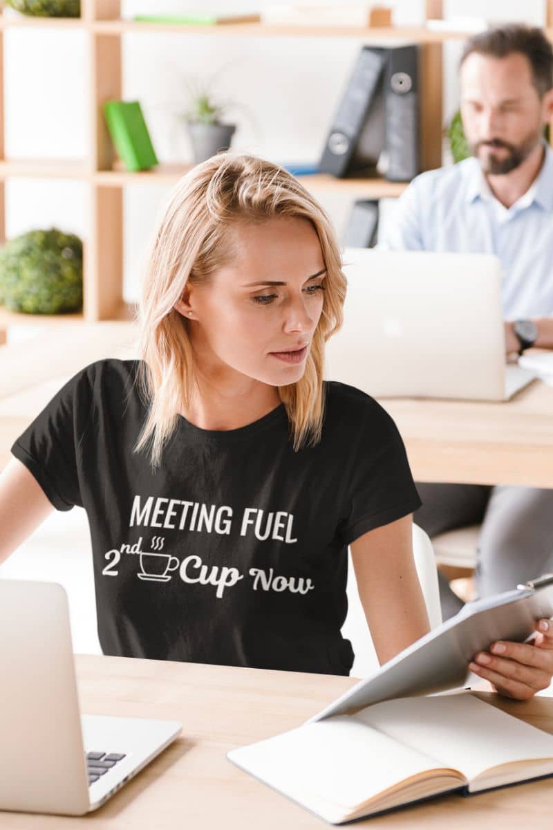 funny coffee shirt with a woman in a black t-shirt with text 'Meeting Fuel: 2nd Cup Now', sitting at a desk in an office setting.