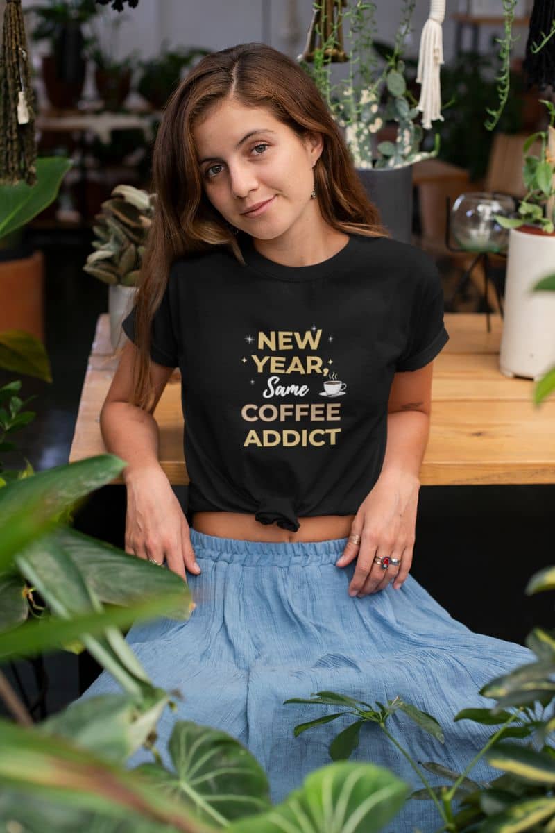 funny coffee shirt with a woman wearing a black t-shirt with 'New Year, Same Coffee Addict' text in a casual setting with plants.
