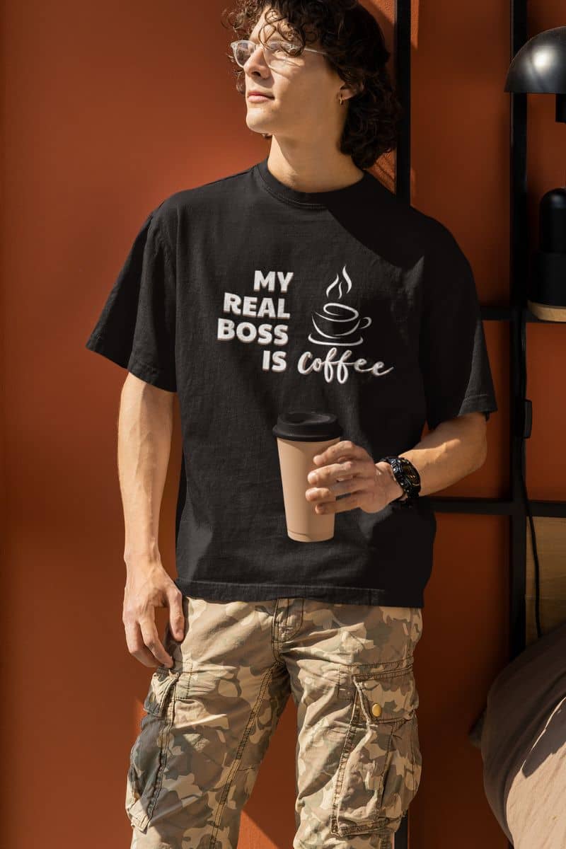 funny coffee t-shirts with a person wearing a black t-shirt with 'My real boss is coffee' text, holding a coffee cup against an orange wall.