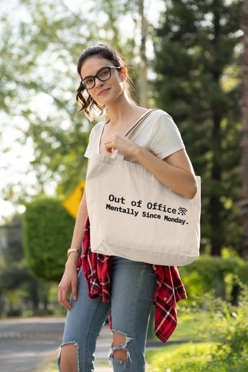 A woman standing outdoors holding a tote bag with the humorous text 'Out of Office. Mentally Since Monday' – a funny corporate life bag.