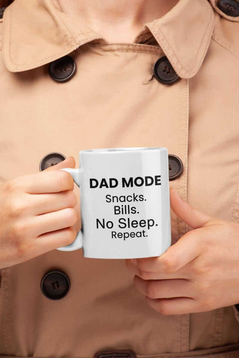 funny dad mug with a person holding a mug with text 'DAD MODE: Snacks.  Bills. No Sleep. Repeat.' in a beige coat.